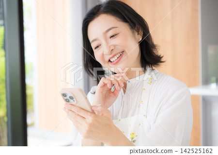 一位五十多歲的女性在家中看著智慧型手機，臉上露出笑容，她一邊做家務，一邊享受著網路和社群媒體帶來的樂趣。 142525162
