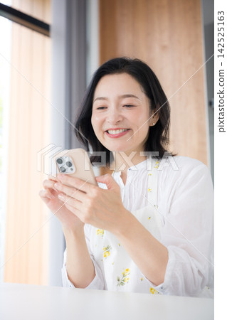 A woman in her 50s smiles while looking at her smartphone at home, reflecting a lifestyle that involves enjoying the internet and social media in between household chores. A woman in her 50s smiles while looking at her smartphone at home, reflecting a lifestyle that involves enjoying the internet and social media in between household chores. 142525163