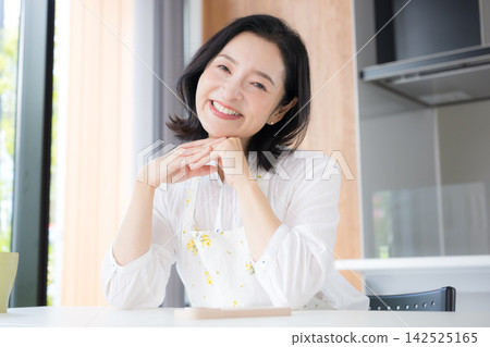 一位中年婦女的特寫鏡頭,她可能是 50 多歲的家庭主婦,穿著圍裙,在自家廚房微笑。 一位中年婦女的特寫鏡頭,她可能是 50 多歲的家庭主婦,穿著圍裙,在自家廚房微笑。 142525165