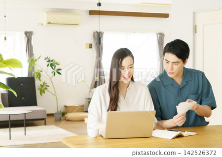 A young Japanese couple at their home table, keeping track of their household expenses on a computer while looking at receipts. 142526372