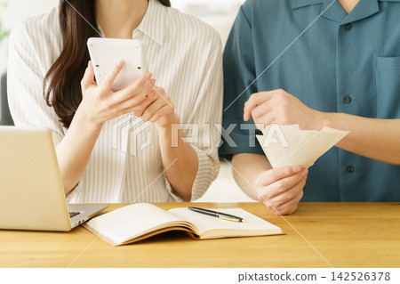 A close-up of the hands of a young Japanese couple sitting at a table in their home, holding a receipt and a calculator. 142526378