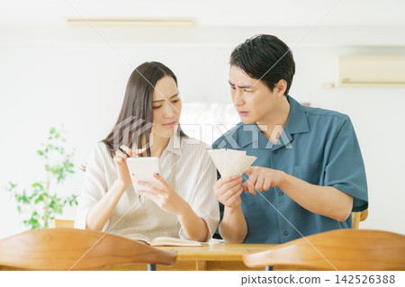 A young Japanese couple is sitting at their table at home, looking at receipts and a calculator, deep in thought. A young Japanese couple is sitting at their table at home, looking at receipts and a calculator, deep in thought. 142526388