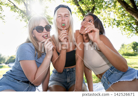 Three young female friends eating lollipops in sunny park Three young female friends eating lollipops in sunny park 142526671