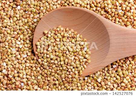 Top view of raw buckwheat groats spread across a surface with a wooden spoon. Uncooked triangular grains Top view of raw buckwheat groats spread across a surface with a wooden spoon. Uncooked triangular grains 142527076