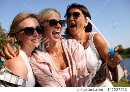 Three happy female friends laughing and blowing soap bubbles together outdoors Three happy female friends laughing and blowing soap bubbles together outdoors 142527722