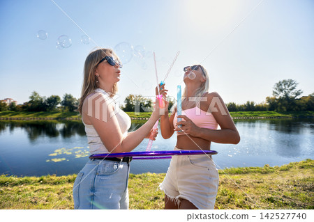 Two young women blowing soap bubbles and playing with hula hoop by lake on sunny day Two young women blowing soap bubbles and playing with hula hoop by lake on sunny day 142527740