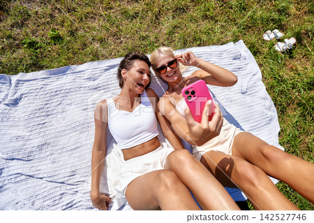 Two happy women taking selfie photo with smartphone while relaxing on grass in park Two happy women taking selfie photo with smartphone while relaxing on grass in park 142527746