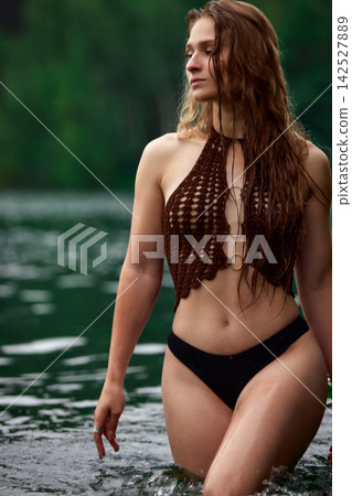 Serene woman in brown crochet bikini top wading in calm green lake Serene woman in brown crochet bikini top wading in calm green lake 142527889