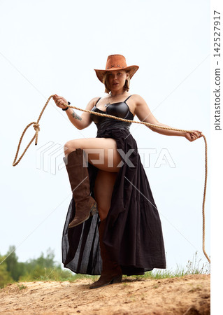 Powerful cowgirl in black dress and cowboy hat holding lasso on sandy hill 142527917