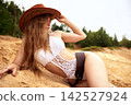 Woman in cowboy hat and white crochet swimsuit lying on sandy hill 142527924