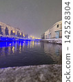 A canal on a snowy night and a cityscape illuminated in blue lights. 142528304
