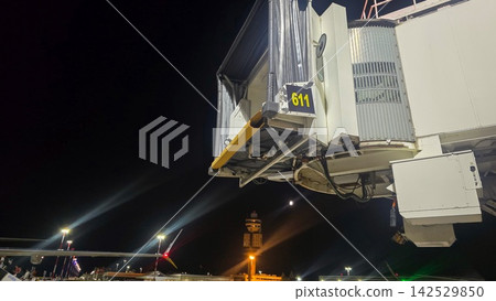 Airport jet bridge and boarding gate stand illuminated at night beside an aircraft. Concepts: air travel, aviation infrastructure, boarding, transportation, tourism, nighttime operations. Airport jet bridge and boarding gate stand illuminated at night beside an aircraft. Concepts: air travel, aviation infrastructure, boarding, transportation, tourism, nighttime operations. 142529850