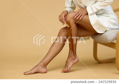 Woman applies wax to lower leg while sitting in armchair Woman applies wax to lower leg while sitting in armchair 142529930