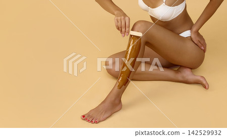 Woman performs leg waxing while seated on beige floor Woman performs leg waxing while seated on beige floor 142529932
