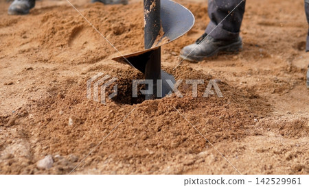 Close view shows metal auger boring into loose sandy soil while worker boots stand nearby, with auger, drilling, excavation, groundwork detail, showing construction, soil, sand, earth context Close view shows metal auger boring into loose sandy soil while worker boots stand nearby, with auger, drilling, excavation, groundwork detail, showing construction, soil, sand, earth context 142529961
