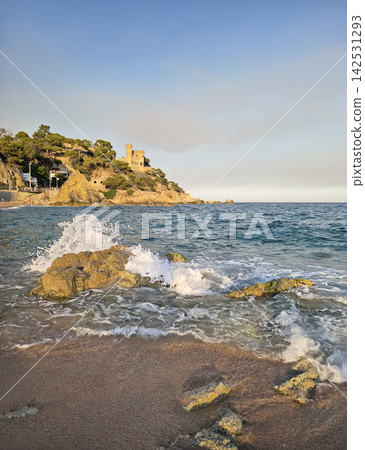 Waves crash against rocks along the shore of Lloret de Mar, Spain, with the iconic Castell d'en Plaja perched on a cliffside above. Warm evening light illuminates the Mediterranean coast in Catalonia Waves crash against rocks along the shore of Lloret de Mar, Spain, with the iconic Castell d'en Plaja perched on a cliffside above. Warm evening light illuminates the Mediterranean coast in Catalonia 142531293