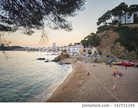 Lloret de Mar, Spain - July 04, 2026: Dusk settles over the sandy beach at Sa Caleta. Gentle Mediterranean waves lap the shore near rocky cliffs and pine trees while tourists relax on the resort coast Lloret de Mar, Spain - July 04, 2026: Dusk settles over the sandy beach at Sa Caleta. Gentle Mediterranean waves lap the shore near rocky cliffs and pine trees while tourists relax on the resort coast 142531294