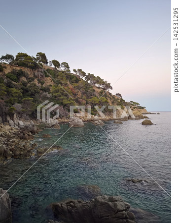 Twilight falls over the rugged rocky coastline of Lloret de Mar, Spain. Calm turquoise Mediterranean waters lap against stone outcrops below steep cliffs covered in pine trees Twilight falls over the rugged rocky coastline of Lloret de Mar, Spain. Calm turquoise Mediterranean waters lap against stone outcrops below steep cliffs covered in pine trees 142531295