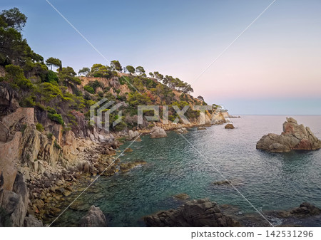 Twilight falls over the rugged rocky coastline of Lloret de Mar, Spain. Calm turquoise Mediterranean waters lap against stone outcrops below steep cliffs covered in pine trees Twilight falls over the rugged rocky coastline of Lloret de Mar, Spain. Calm turquoise Mediterranean waters lap against stone outcrops below steep cliffs covered in pine trees 142531296