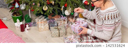 A woman aged 52 sits on the floor next to a decorated Christmas tree. She opens gifts on Christmas Day while enjoying the holiday with family members in the background A woman aged 52 sits on the floor next to a decorated Christmas tree. She opens gifts on Christmas Day while enjoying the holiday with family members in the background 142531624