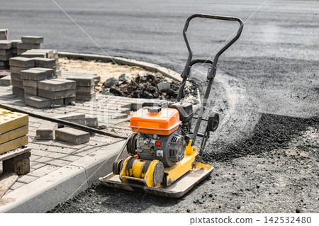 A machine for compacting surface materials stands ready beside bricks laid out for a road work project. The area shows signs of construction activity 142532480