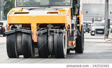 A heavy-duty roller works on a city street, compacting the asphalt as cars pass by. The machine effectively prepares the surface 142532481