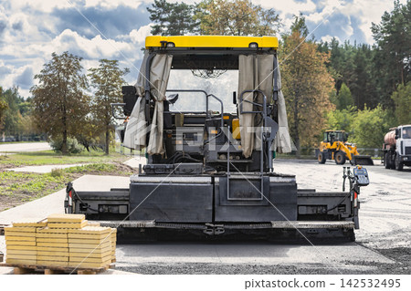 An asphalt paver stands on the roadway, preparing for work. Stacks of materials are placed on the ground near the rear of the machine. Roadway reconstruction An asphalt paver stands on the roadway, preparing for work. Stacks of materials are placed on the ground near the rear of the machine. Roadway reconstruction 142532495