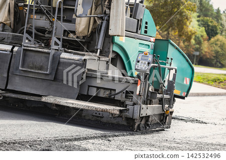 Paving machine works on the road surface, spreading asphalt evenly. The equipment operates steadily, ensuring proper road maintenance in the area Paving machine works on the road surface, spreading asphalt evenly. The equipment operates steadily, ensuring proper road maintenance in the area 142532496