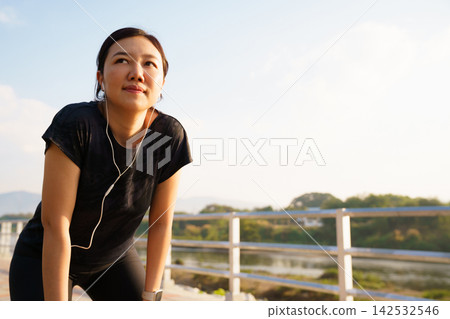 Asian woman resting after running workout on sunny morning Asian woman resting after running workout on sunny morning 142532546