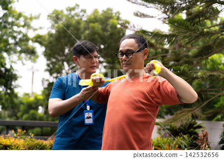 Caregiver helping senior man exercising with resistance band in garden 142532656