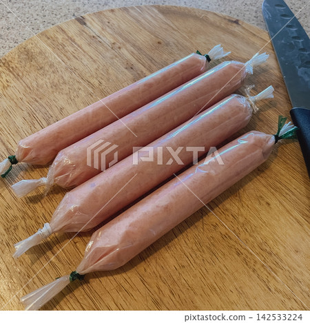 Homemade sausages wrapped in clear plastic on a wooden cutting board with a knife positioned nearby, showcasing a fresh and appetizing presentation of artisanal food preparation Homemade sausages wrapped in clear plastic on a wooden cutting board with a knife positioned nearby, showcasing a fresh and appetizing presentation of artisanal food preparation 142533224