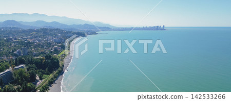 Aerial view of Batumi coastline featuring sandy beach, turquoise sea, and distant mountains under clear blue sky, showcasing urban development along the shoreline 142533266