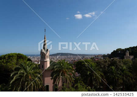 Decorative church spire stands among Mediterranean palms while Barcelona spreads across hillside toward coastline. Bright daylight, open sky, and visible city texture support travel planning imagery. Decorative church spire stands among Mediterranean palms while Barcelona spreads across hillside toward coastline. Bright daylight, open sky, and visible city texture support travel planning imagery. 142533425
