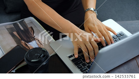 Hands typing on laptop beside magazine, smartphone and coffee cup at desk, showing focused work on creative project, online communication, planning and remote productivity in modern workspace 142533761