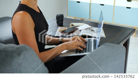 Woman typing on laptop beside notebook, smartphone, coffee cup, and open magazine while sitting on sofa in bright modern home interior with relaxed workspace setup and daily planning Woman typing on laptop beside notebook, smartphone, coffee cup, and open magazine while sitting on sofa in bright modern home interior with relaxed workspace setup and daily planning 142533762