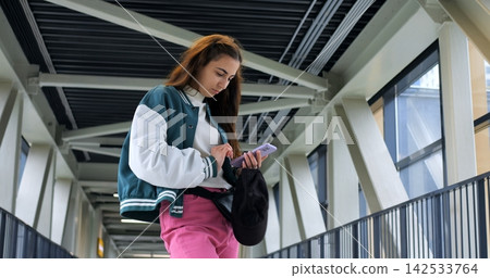 Young woman searching bag for ticket on walkway, hands inside black shoulder bag while checking phone, practical transit moment with structural beams and railings in background, focused movement Young woman searching bag for ticket on walkway, hands inside black shoulder bag while checking phone, practical transit moment with structural beams and railings in background, focused movement 142533764