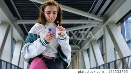 Woman on pedestrian bridge checking phone while holding pink lollipop and wearing varsity jacket, pausing midwalk under modern steel walkway, absorbed in message and notifications. Woman on pedestrian bridge checking phone while holding pink lollipop and wearing varsity jacket, pausing midwalk under modern steel walkway, absorbed in message and notifications. 142533765