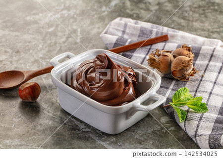 Rich chocolate hazelnut spread in white ceramic dish with fresh mint and whole hazelnuts on checkered cloth 142534025