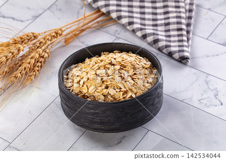 Rolled oats in black ceramic bowl with wheat stalks and checkered cloth on marble surface Rolled oats in black ceramic bowl with wheat stalks and checkered cloth on marble surface 142534044