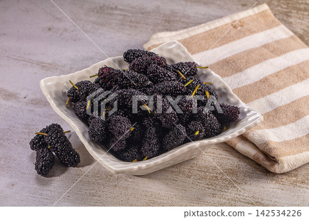 Fresh dark mulberries in white ceramic bowl on rustic surface with striped cloth Fresh dark mulberries in white ceramic bowl on rustic surface with striped cloth 142534226