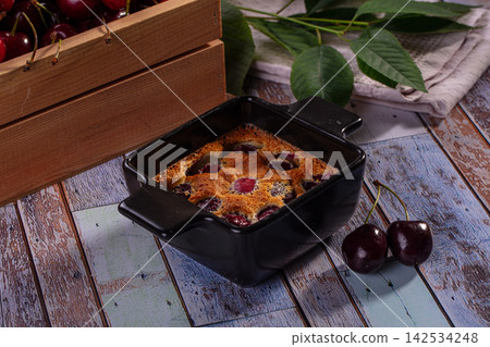 Baked cherry clafoutis in black ceramic dish with fresh cherries and green leaves on rustic wooden table Baked cherry clafoutis in black ceramic dish with fresh cherries and green leaves on rustic wooden table 142534248