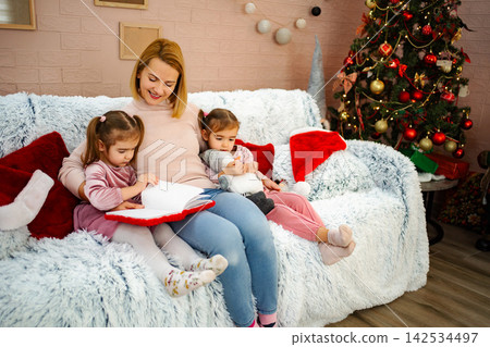 Mother reading book to daughters by Christmas tree Mother reading book to daughters by Christmas tree 142534497