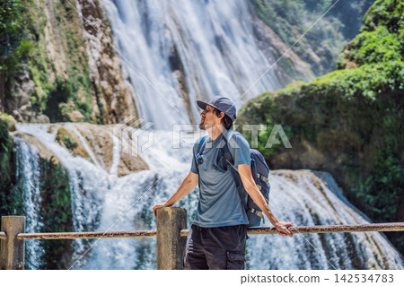 Man traveler standing near El Chiflon waterfall with a backpack in Chiapas Mexico with wooden railings tropical forest clear blue water nature travel and looking at the waterfall. Man traveler standing near El Chiflon waterfall with a backpack in Chiapas Mexico with wooden railings tropical forest clear blue water nature travel and looking at the waterfall. 142534783