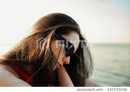 Woman in sunglasses leans toward the sea at golden hour, appearing calm and introspective as warm light highlights her hair and profile, capturing solitude, freedom, relaxation, and a peaceful escape. Woman in sunglasses leans toward the sea at golden hour, appearing calm and introspective as warm light highlights her hair and profile, capturing solitude, freedom, relaxation, and a peaceful escape. 142535036
