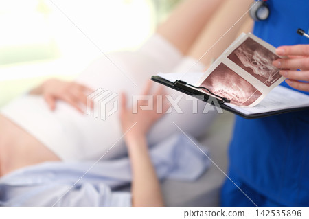 Healthcare professional reviewing ultrasound scan images beside pregnant patient in hospital room during prenatal consultation, with focus on medical discussion, patient care, and diagnostic review Healthcare professional reviewing ultrasound scan images beside pregnant patient in hospital room during prenatal consultation, with focus on medical discussion, patient care, and diagnostic review 142535896