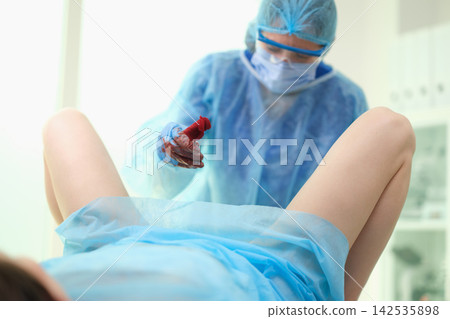 Medical clinician in protective gear performing pelvic examination with patient on procedure table in bright clinic, gloved hand holding bloody swab, visible legs raised for examination Medical clinician in protective gear performing pelvic examination with patient on procedure table in bright clinic, gloved hand holding bloody swab, visible legs raised for examination 142535898