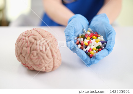 Gloved hands holding colorful capsules beside brain model, illustrating medical research, neurology, mental health treatment, pharmacology, and healthcare concepts in clean clinical setting Gloved hands holding colorful capsules beside brain model, illustrating medical research, neurology, mental health treatment, pharmacology, and healthcare concepts in clean clinical setting 142535901