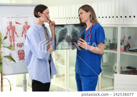 Female doctor in blue scrubs showing chest xray to patient during consultation in bright medical office with anatomy poster, cabinets, and professional clinical atmosphere Female doctor in blue scrubs showing chest xray to patient during consultation in bright medical office with anatomy poster, cabinets, and professional clinical atmosphere 142536077
