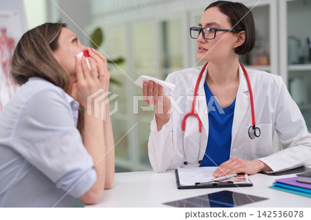 Female doctor in white coat with red stethoscope offering tissue to upset patient during consultation in bright clinic office, showing caring healthcare support and medical communication Female doctor in white coat with red stethoscope offering tissue to upset patient during consultation in bright clinic office, showing caring healthcare support and medical communication 142536078