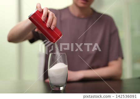 Person pouring white granulated sugar from red container into clear glass on table, close-up indoor scene with blurred background, sweetener concept, portion control, cooking ingredient, diabetes Person pouring white granulated sugar from red container into clear glass on table, close-up indoor scene with blurred background, sweetener concept, portion control, cooking ingredient, diabetes 142536091